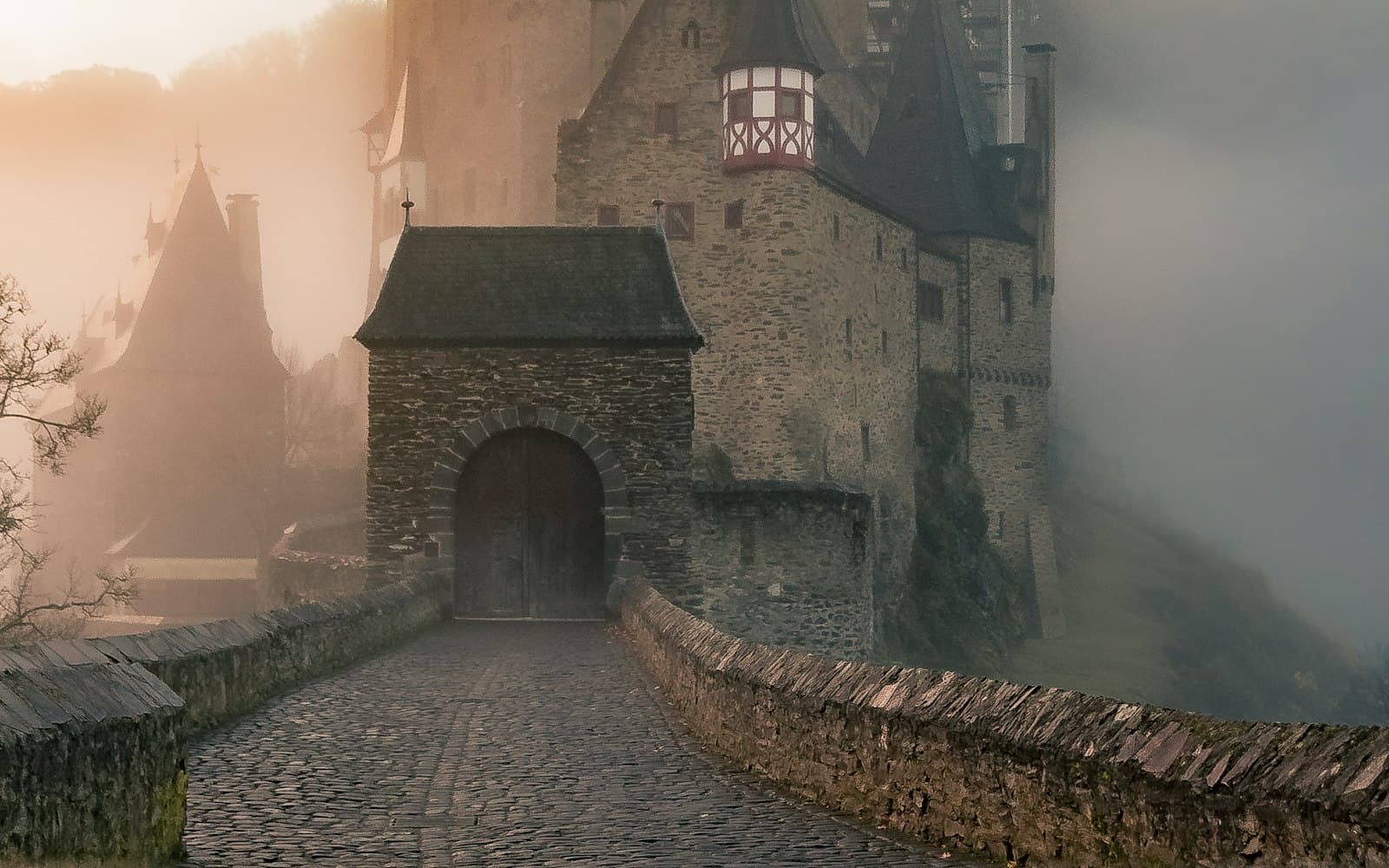 A stone castle gate in heavy fog, with a cobbled bridge leading to wooden doors